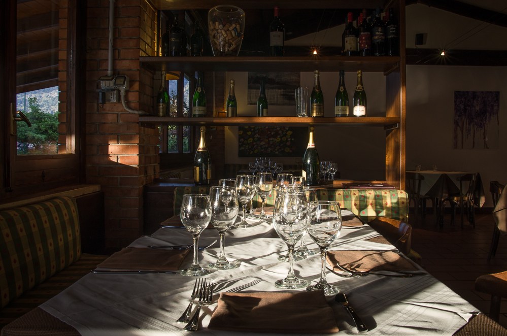 Luce calda di lampada sulle bottiglie allineate sugli scaffali di legno, accanto a un divanetto a righe verdi e crema.